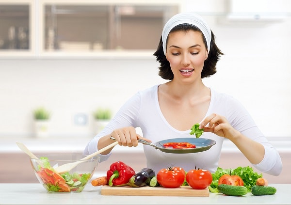 A brunette woman wearing a white hair band and a white shirt in the kitchen, making a salad